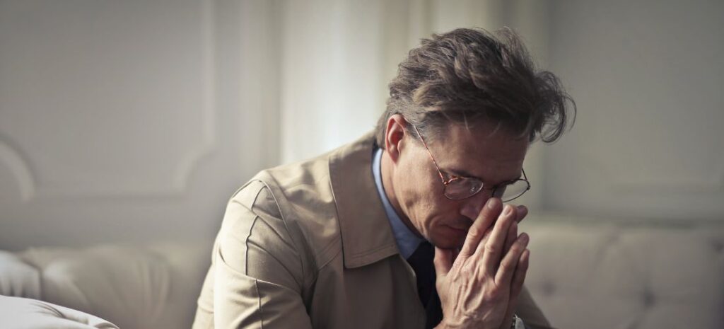 A man in a beige jacket and glasses sits with his hands clasped, looking down in a contemplative or worried state.