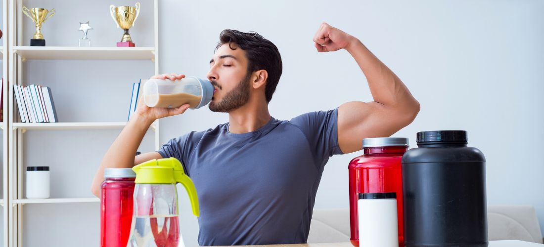 A man drinking a protein shake and flexing his bicep, surrounded by various protein powder containers as a way to gain weight.